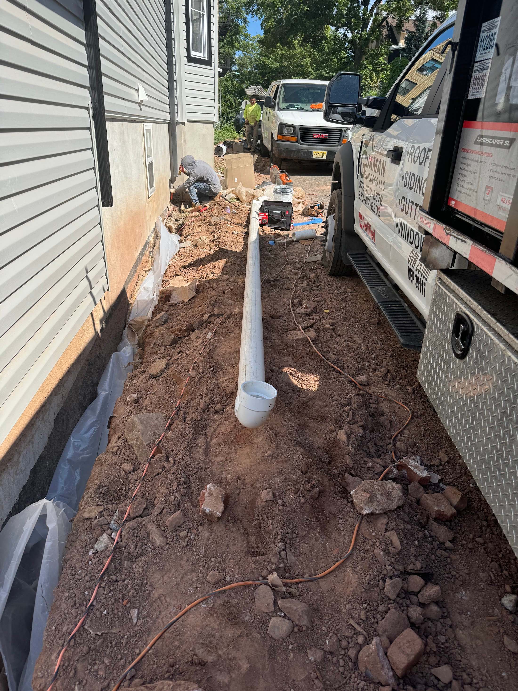 A white downspout drain pipe laid in a fresh trench along a house foundation, beside the Guaman truck