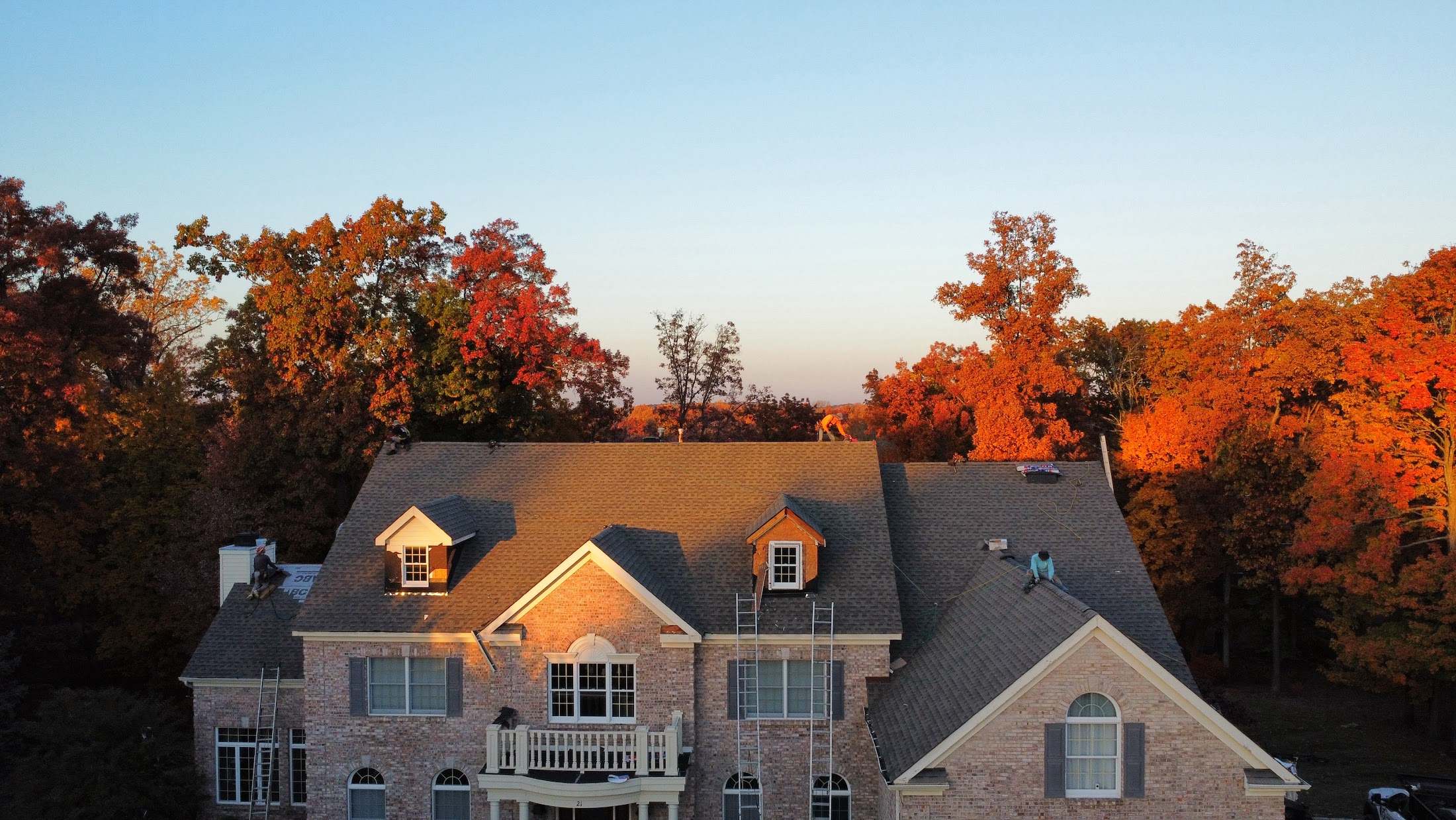 Brick colonial with new shingles on the roof at sunset, two crew members still working up top