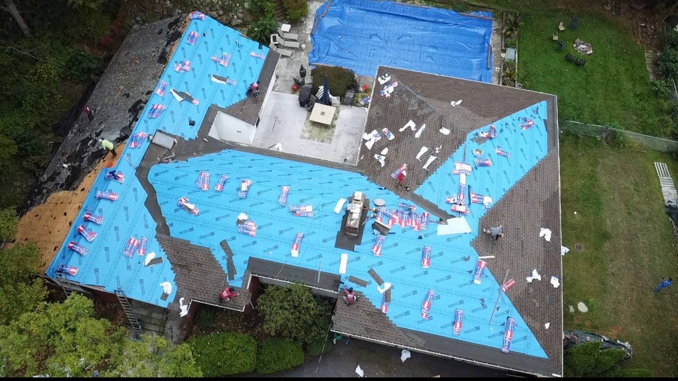 Drone view of a large house mid tear-off, blue underlayment going down over the deck and shingle bundles staged across the roof