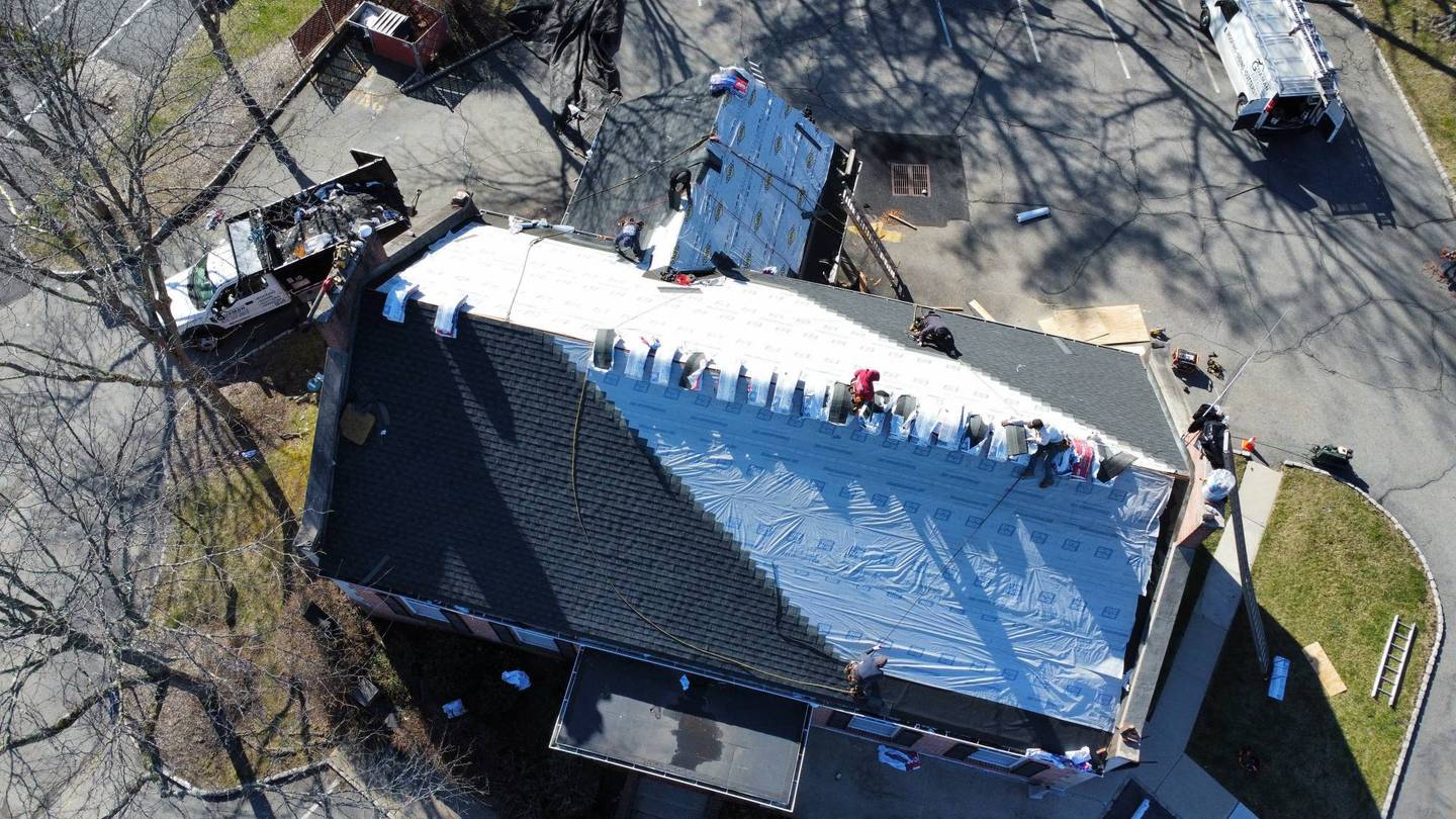 Drone view of a crew re-roofing a long brick building: new underlayment on one side, the company truck and van parked beside it