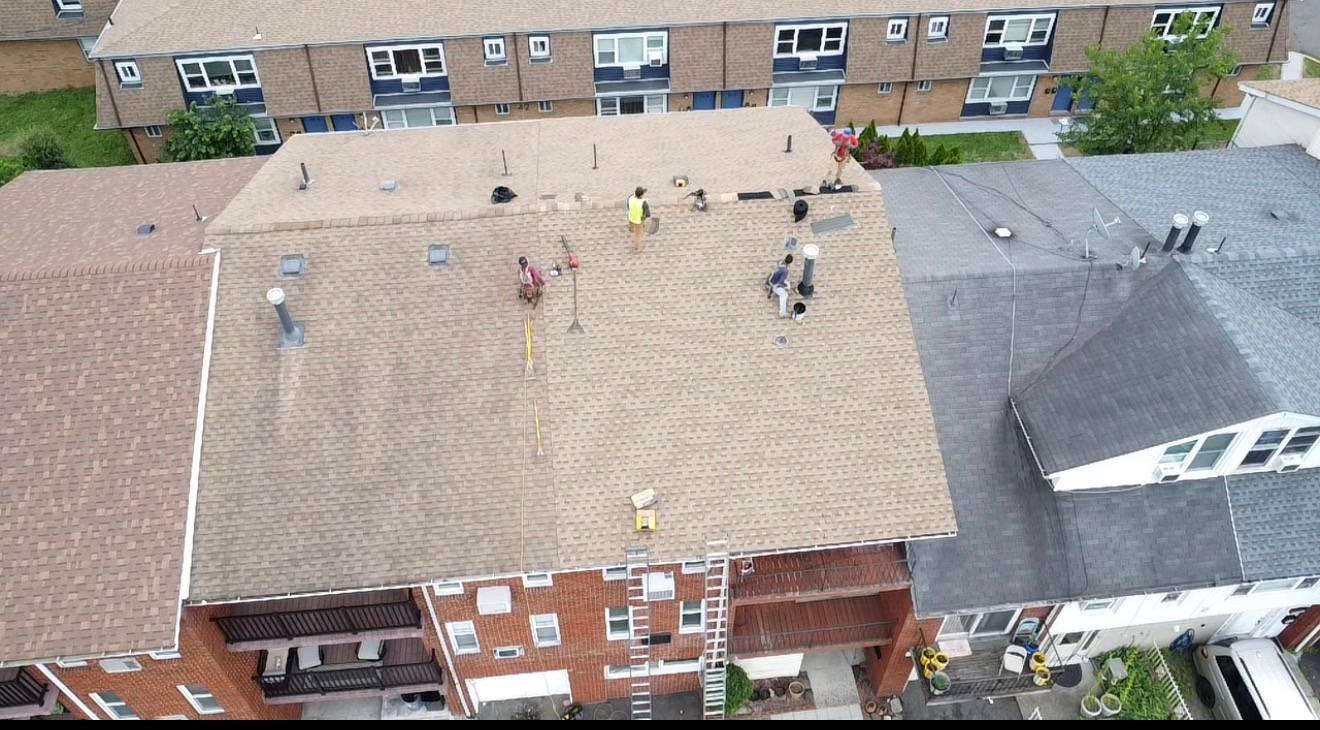 Drone view of roofers working on a tan shingle roof of an attached brick house, ladders against the wall