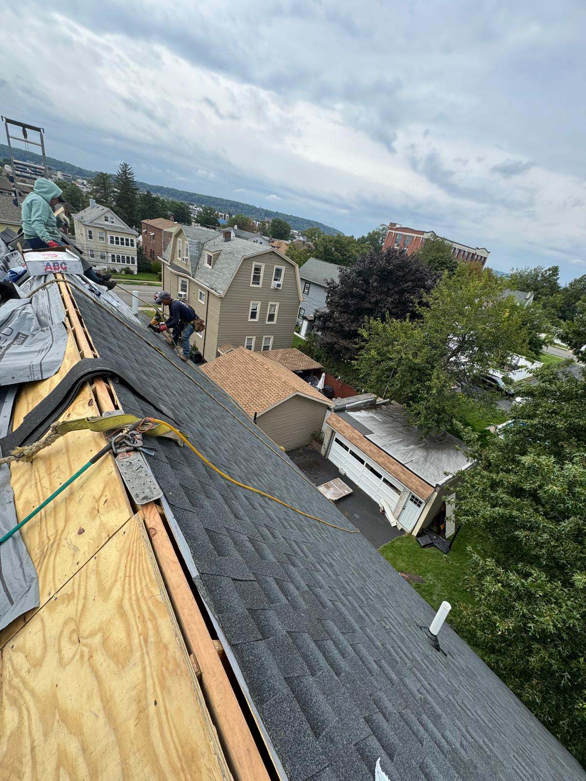 Two roofers tied off with safety lines installing dark shingles on a steep roof