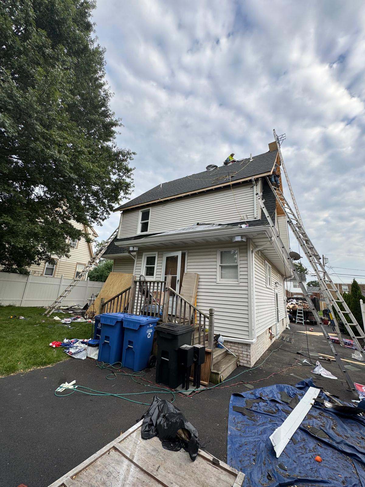 Back of a two-story house with ladders against it while roofers install dark shingles
