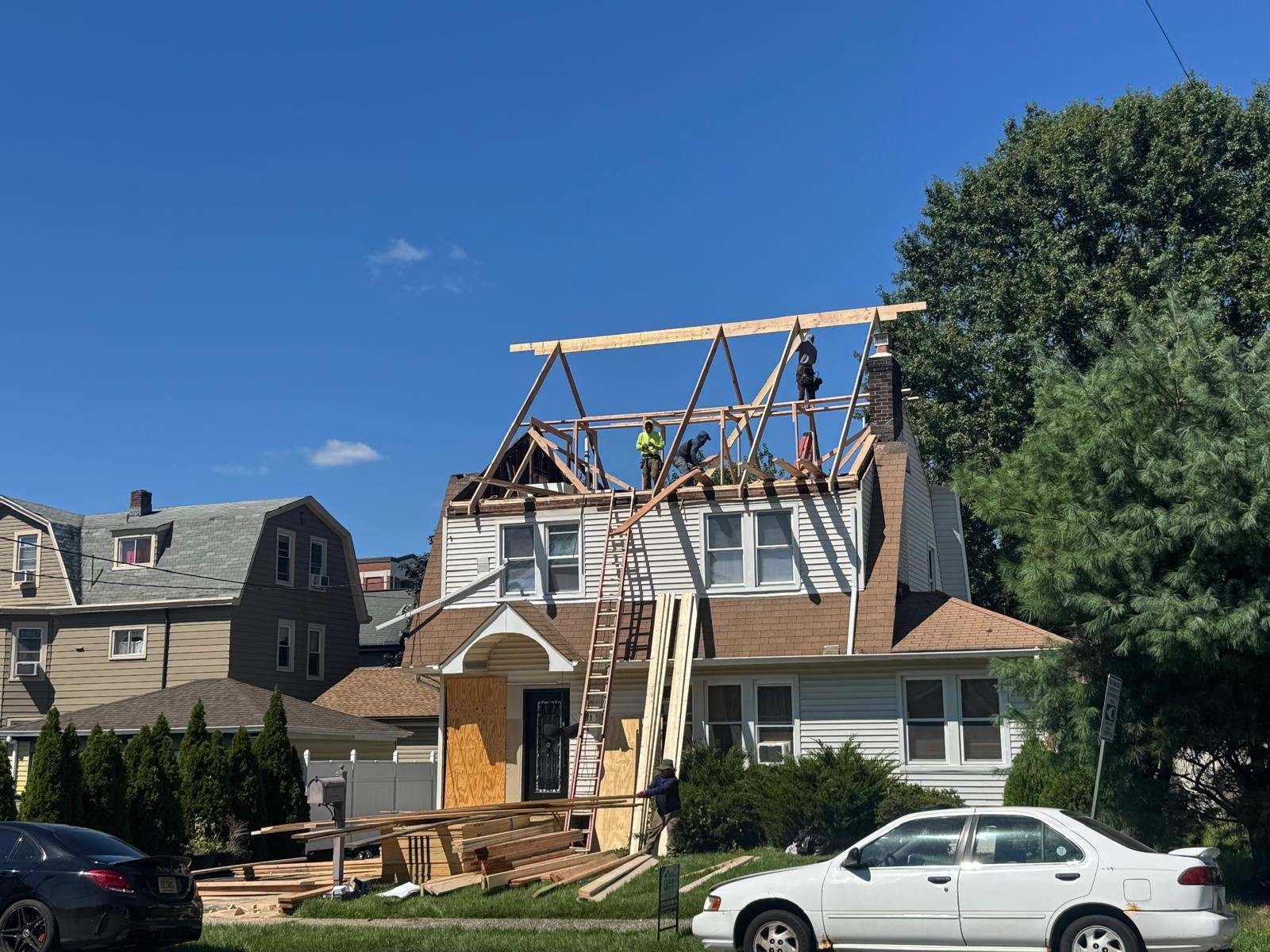 Crew framing a new roof with wood rafters on top of a two-story house, lumber stacked on the lawn