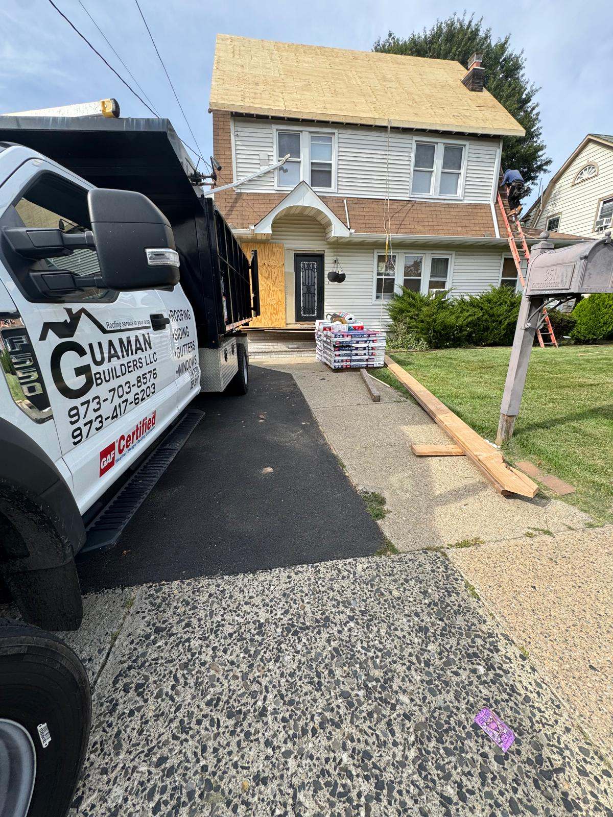 Guaman Builders dump truck in a driveway in front of a house with a new plywood roof deck and shingle bundles on the walkway