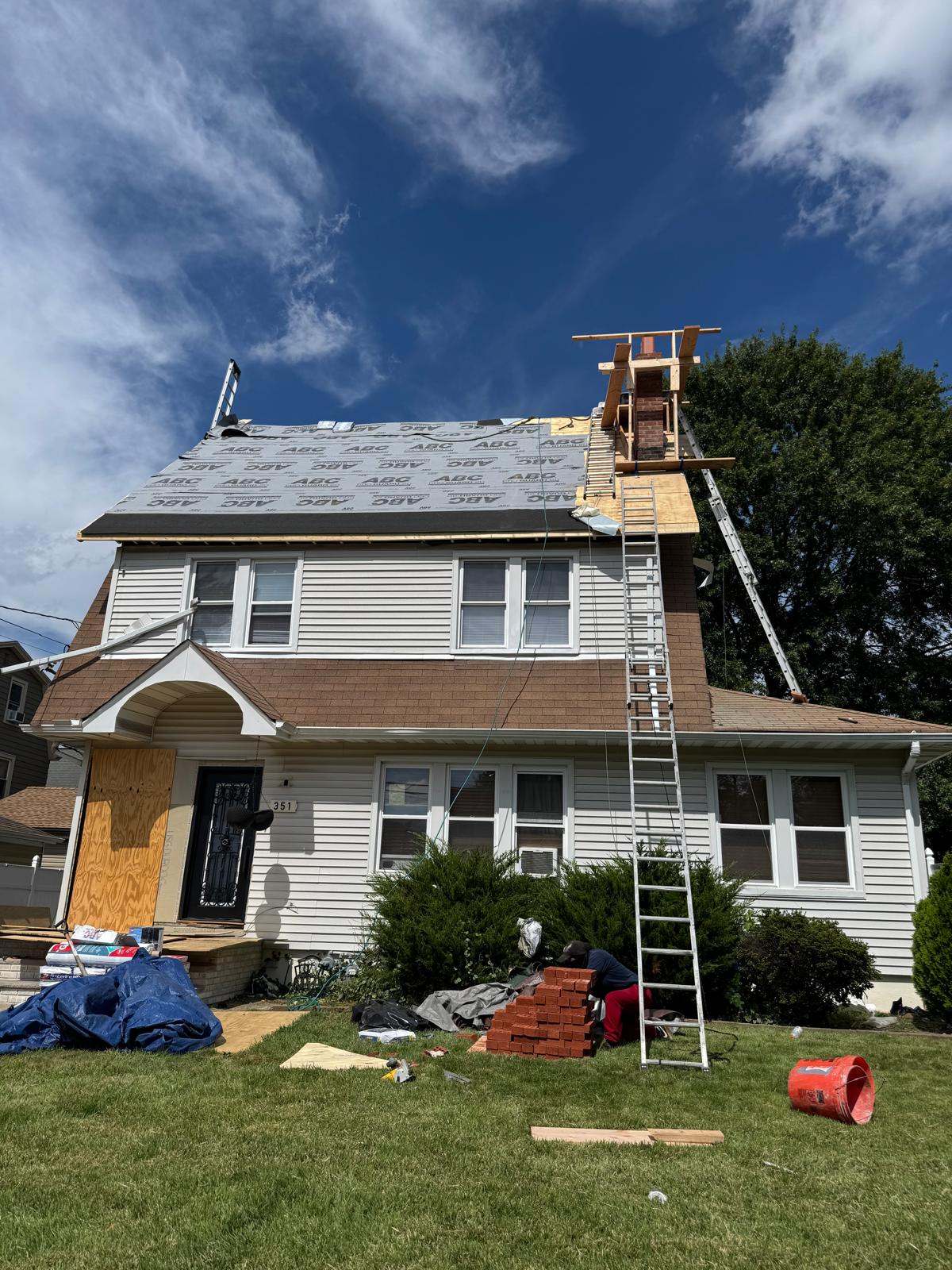 Two-story house with underlayment on the upper roof and wood scaffolding around the chimney, bricks stacked on the lawn