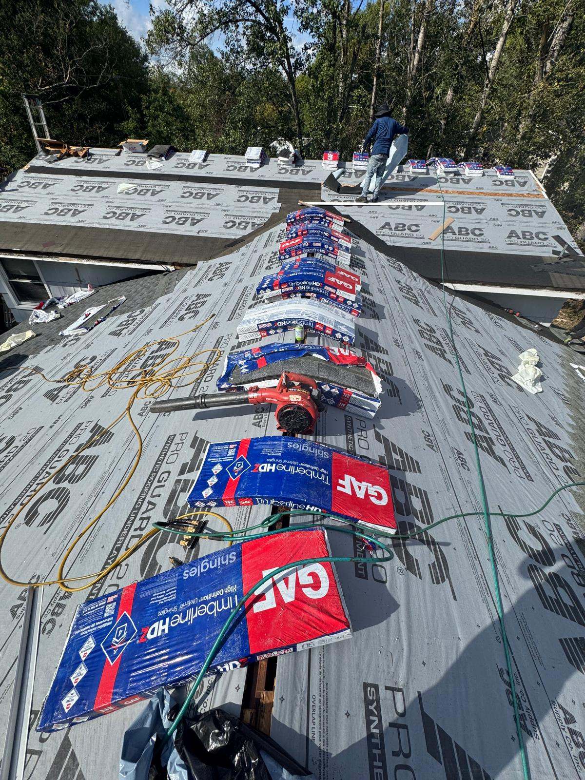 Roof covered in gray synthetic underlayment with GAF Timberline HDZ shingle bundles lined up along the ridge and a roofer working at the top