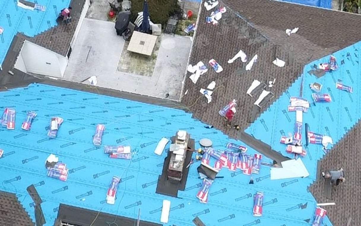 The same house with blue synthetic underlayment across the roof and shingle bundles staged on top